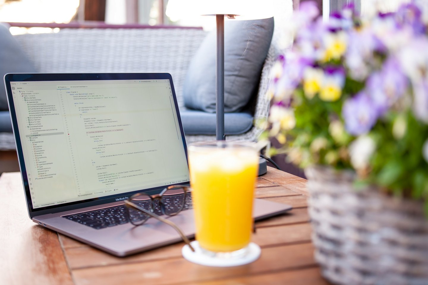 Laptop displaying computer code on a wooden outdoor table with orange juice and flowers.