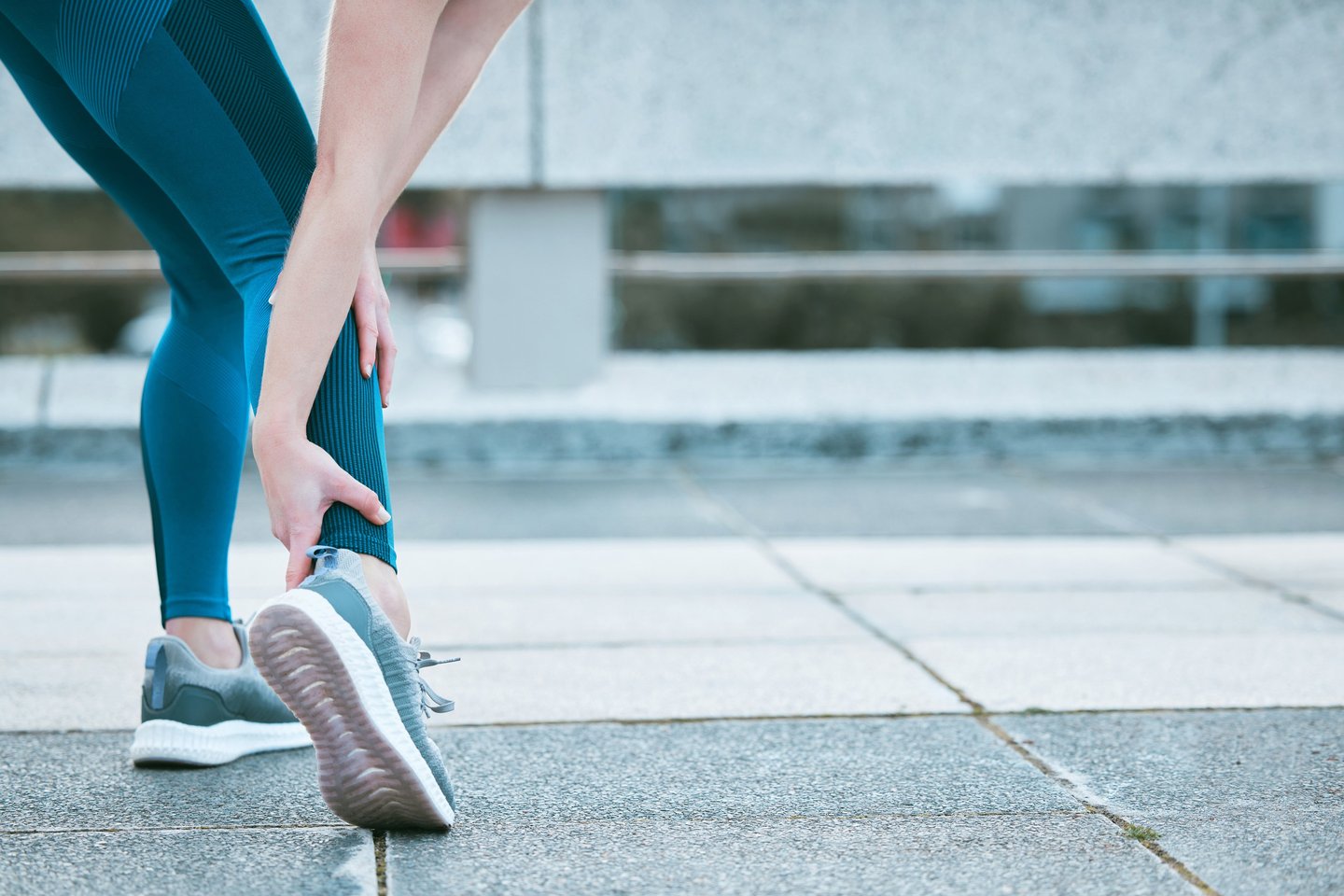 A woman in blue leggings holding her ankle due to a sports injury or sprain while running outdoors.