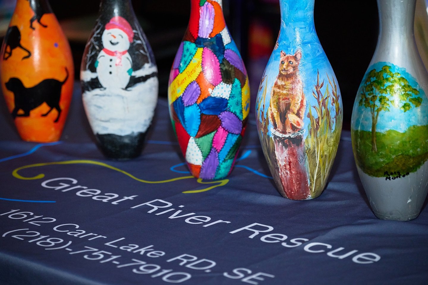 Hand-painted bowling pins featuring art of a cat, snowman, and nature on a Great River Rescue table.