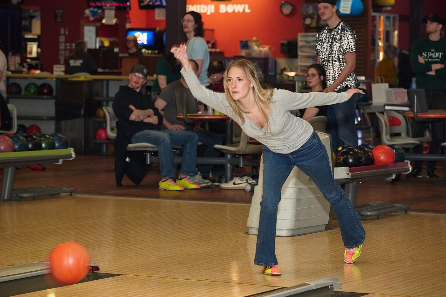 A blonde woman in jeans throws an orange bowling ball down the lane at a local bowling alley.