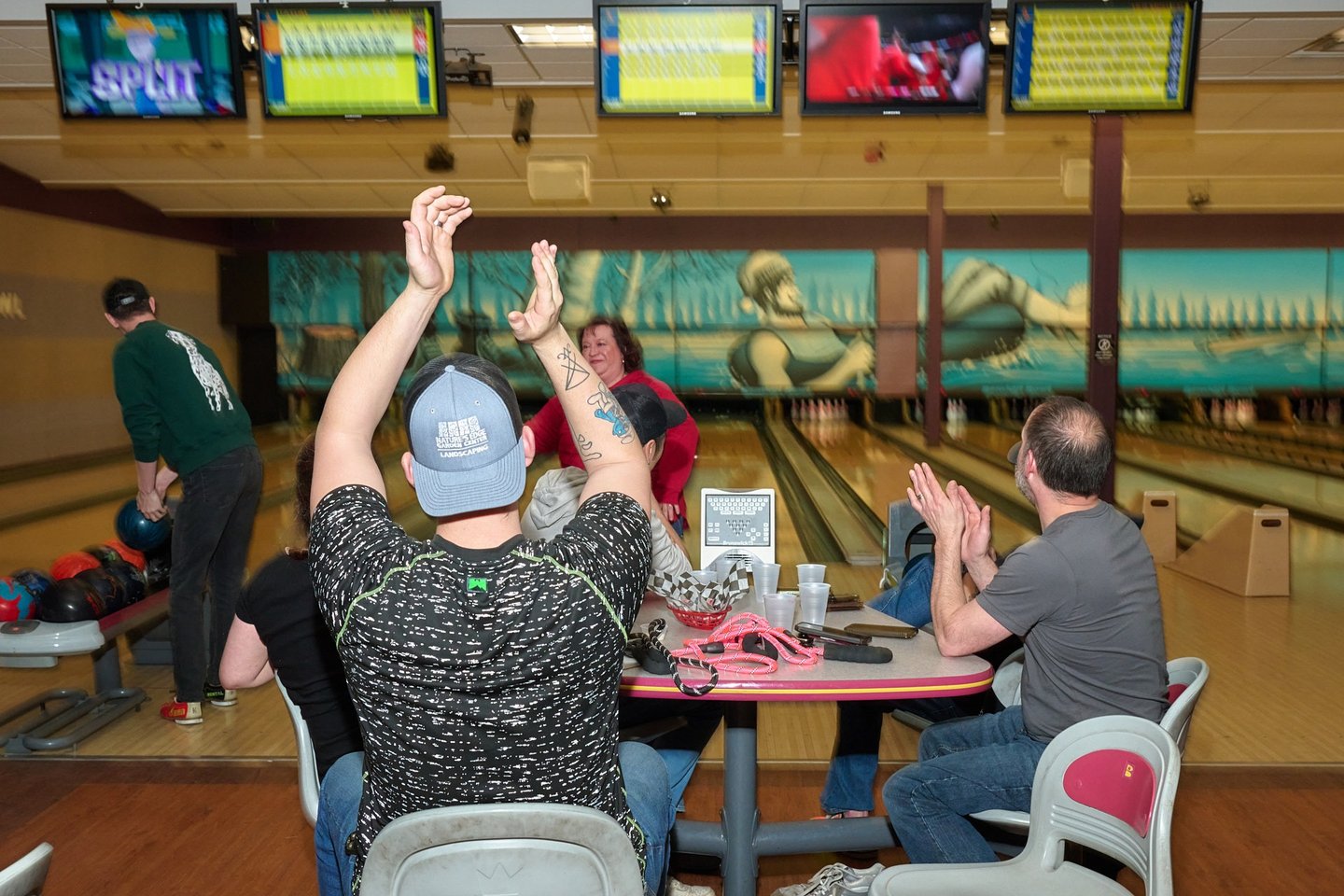 A group of friends celebrating and clapping at a bowling alley with lanes and score monitors.
