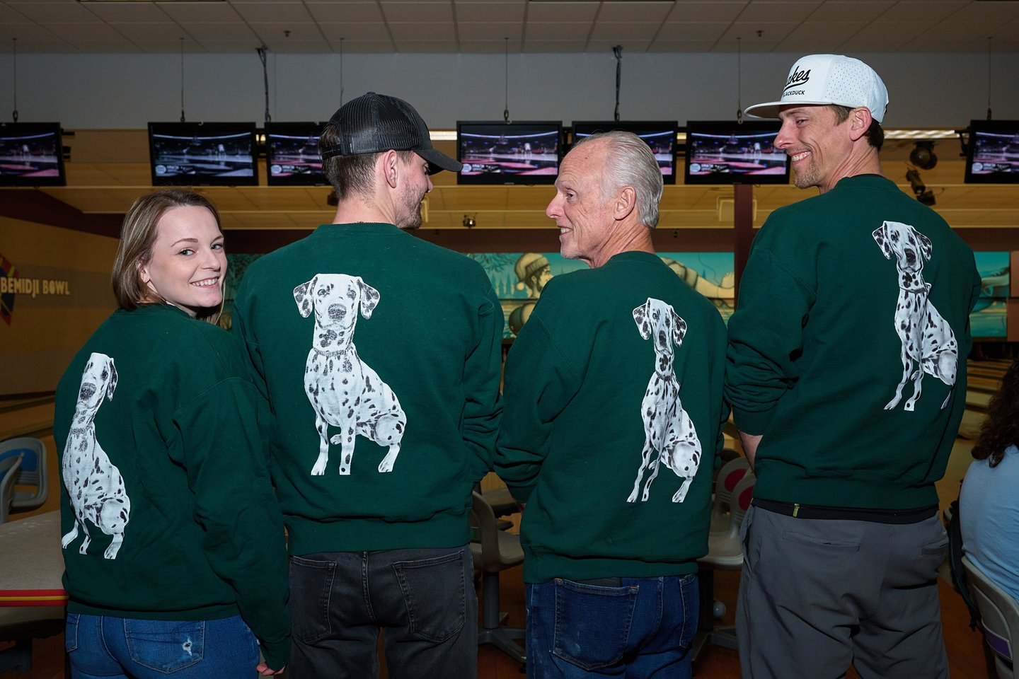 Four smiling people at a bowling alley wearing matching green sweatshirts with Dalmatian dog graphics.