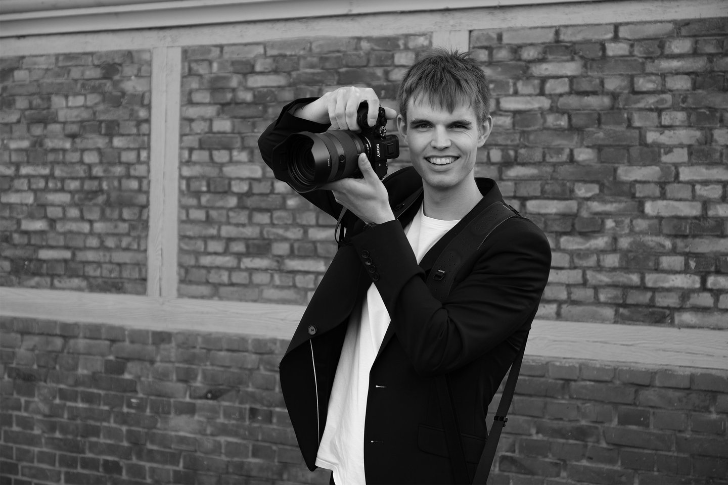 Schwarz-Weiß-Aufnahme von Joel Pingel mit Kamera vor einer Ziegelwand. Eventfotografie: Just Perspectives.