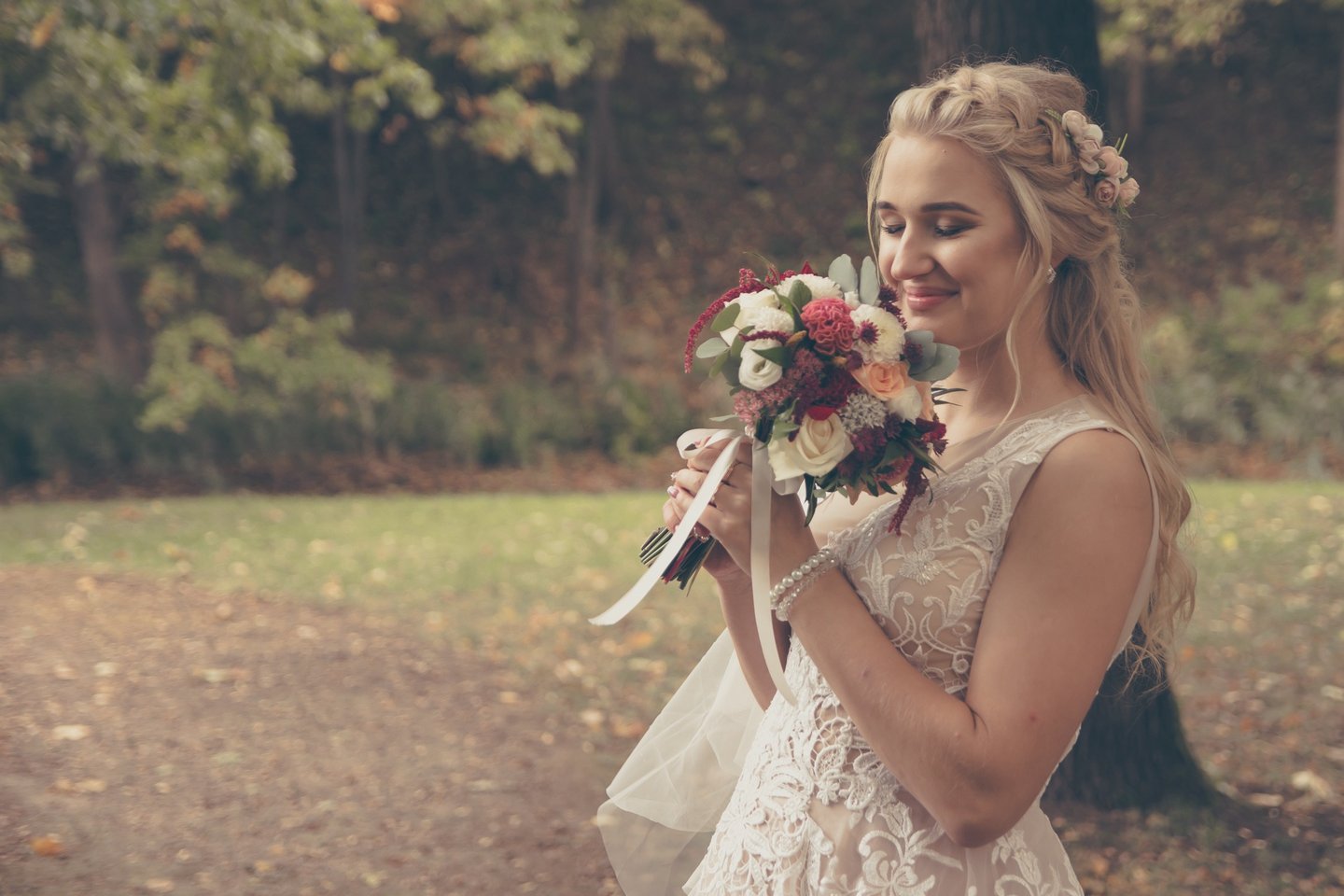 Nuotaka švelniai uodžia savo gėlių puokštę, ant jos rankų perlų apyrankė, romantiška šukuosena