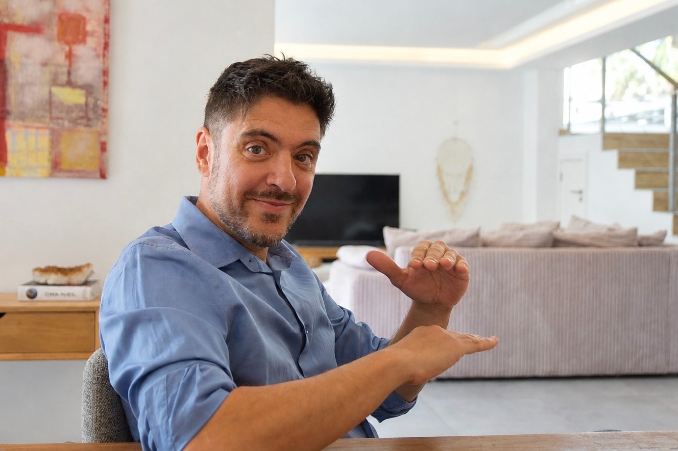 A man in a blue shirt uses hand gestures while sitting in a modern living room setting.