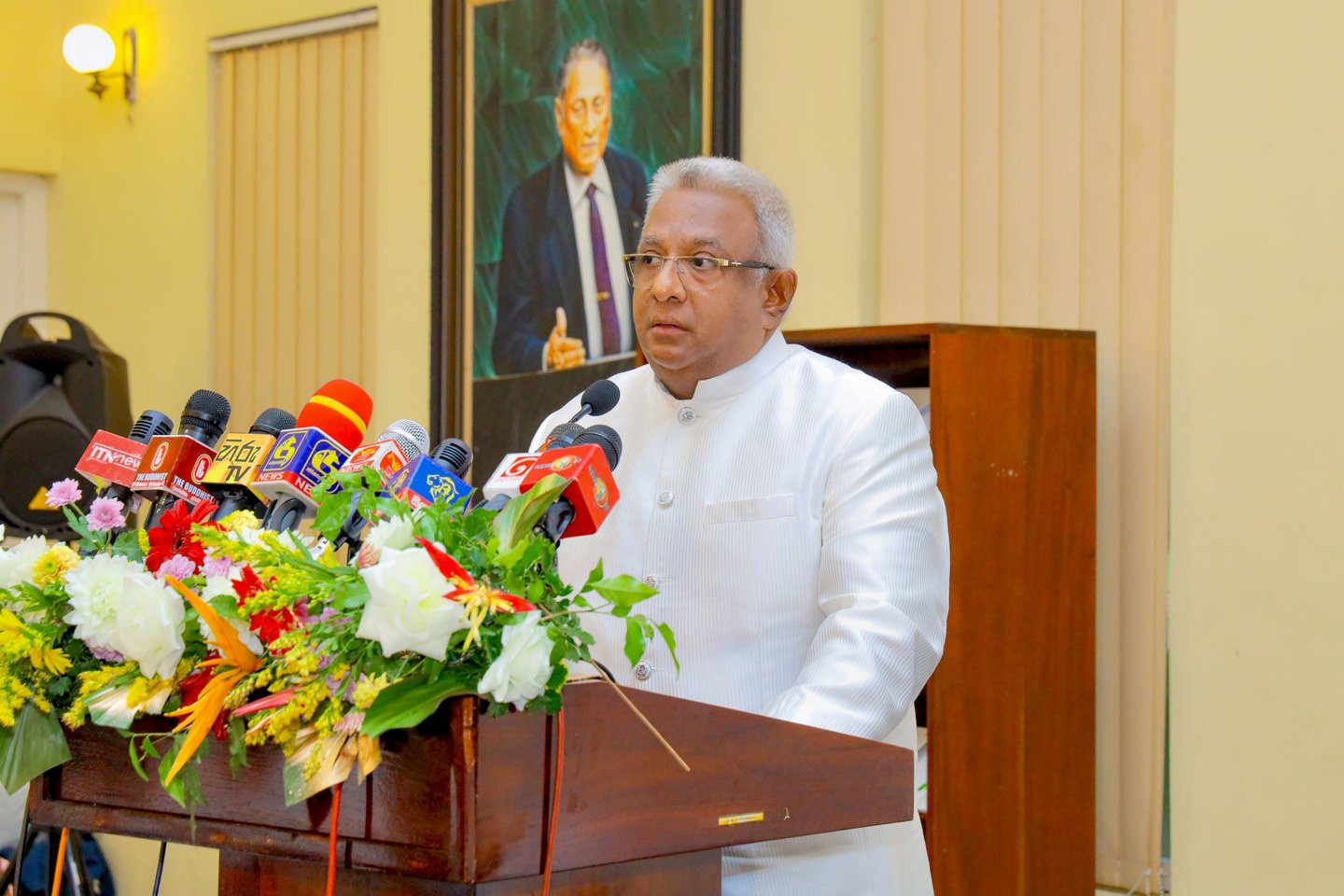 a man in a white suit and glasses speaking into microphones