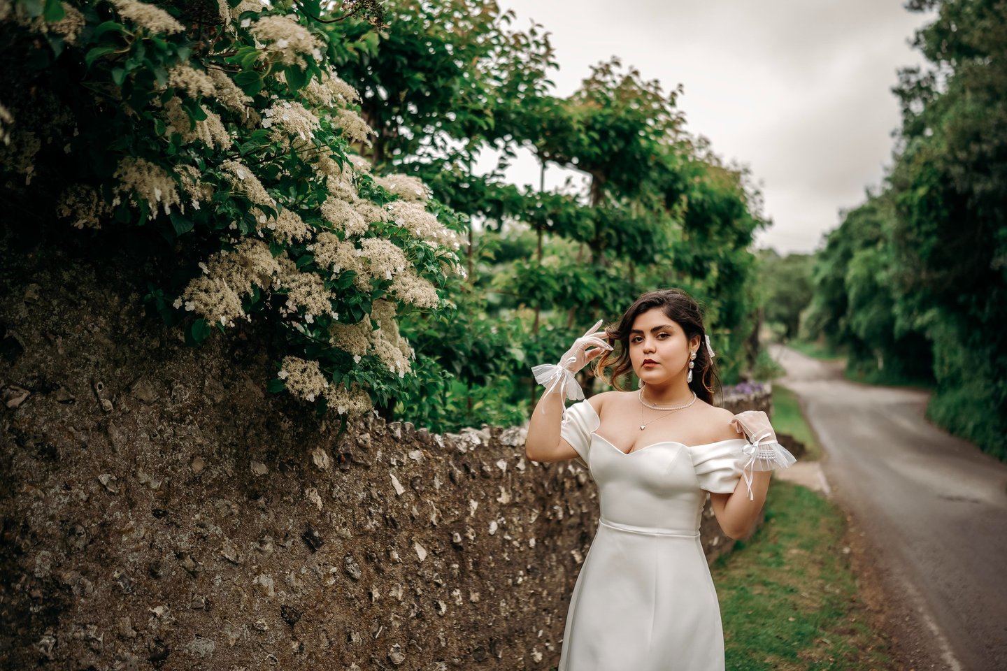 Bride in white dress posing by a stone wall with lush greenery, photographed in Kent.
