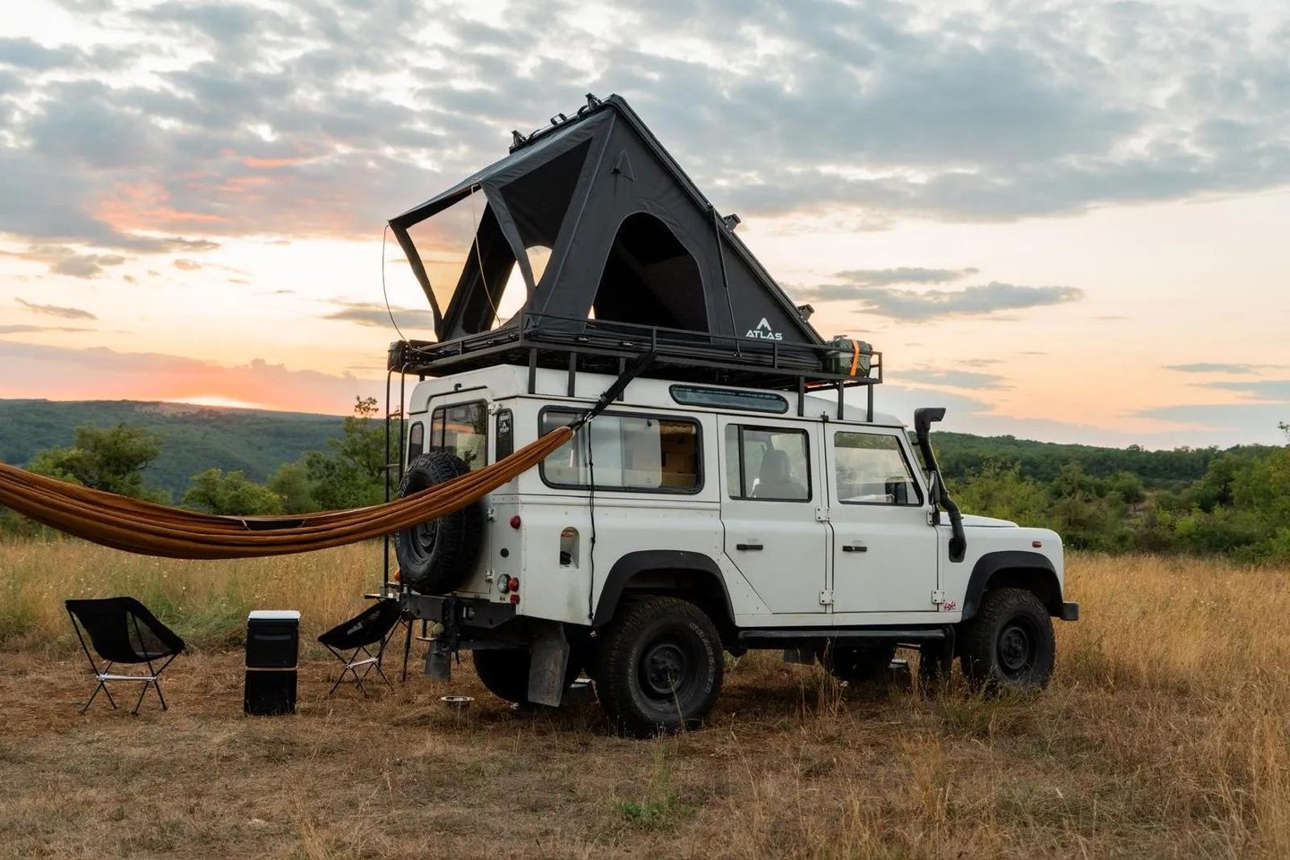 A white Land Rover Defender with a roof top tent and hammock parked in a field at sunset.
