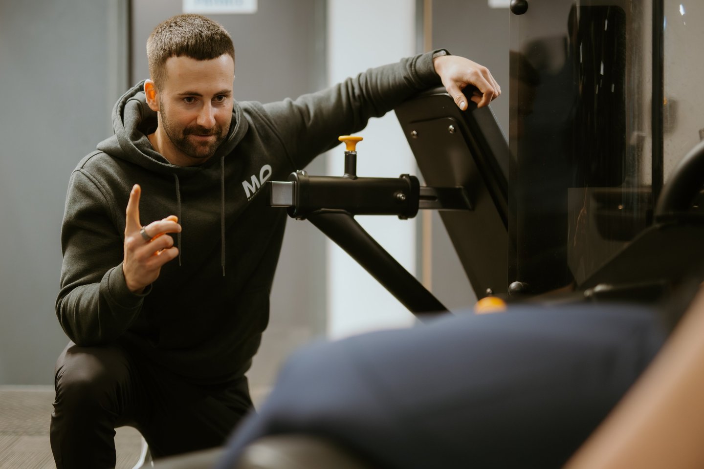 Personal trainer in hoodie coaching client on gym equipment in Leamington Spa fitness centre, focused on technique and su