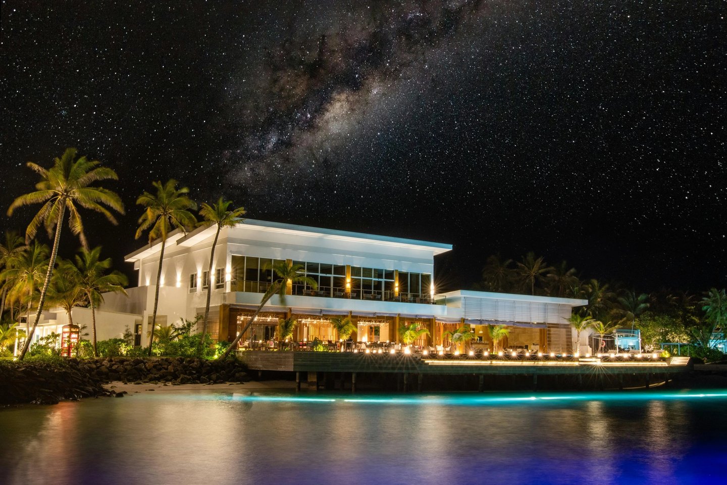 Luxury tropical resort at night featuring the Milky Way galaxy above a lit beachfront restaurant and palm trees.