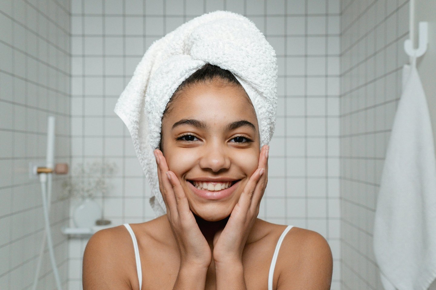A smiling woman with a white towel head wrap shows off her glowing skin during a bathroom skincare routine.