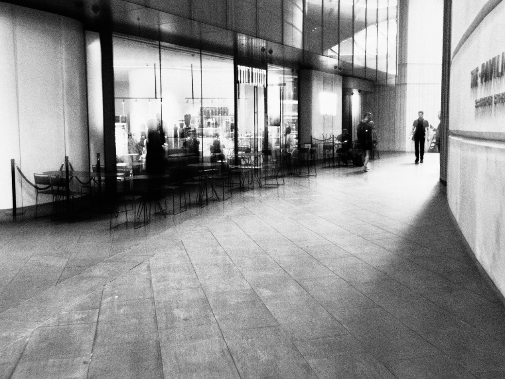 A modern cafe with people moving inside. A lone figure walks on a wide, empty tiled hall, lending a serene urban atmosphere.