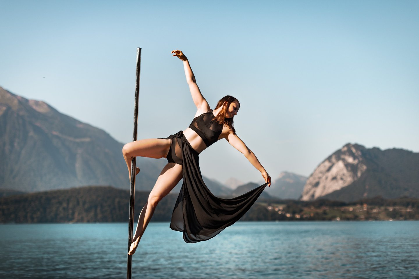 Sabrina Bel Pole doing a cupid pole trick outdoors, with a lake and mountains in the background.