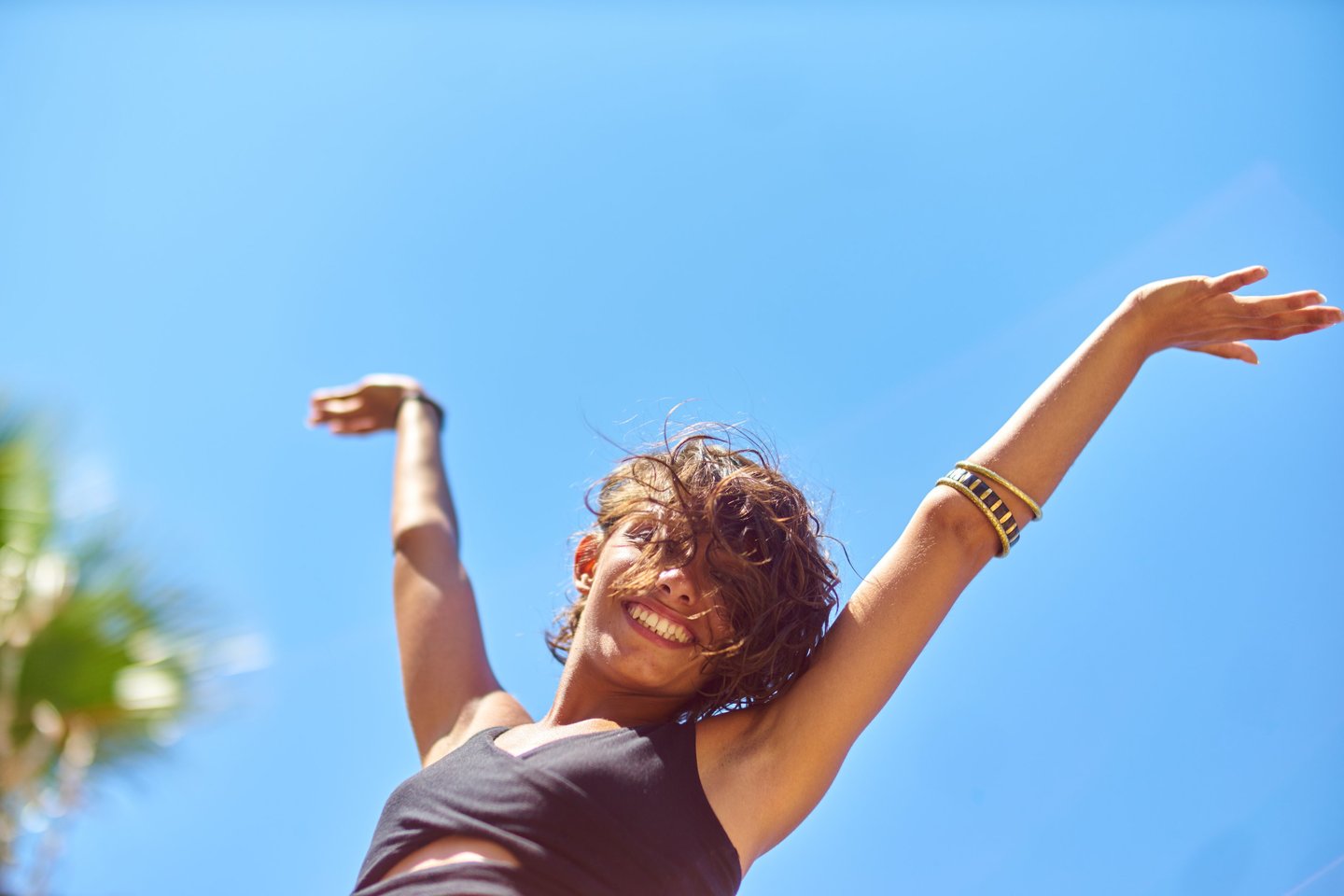 A happy woman with arms raised against a clear blue sky, expressing freedom and joy during a summer vacation.