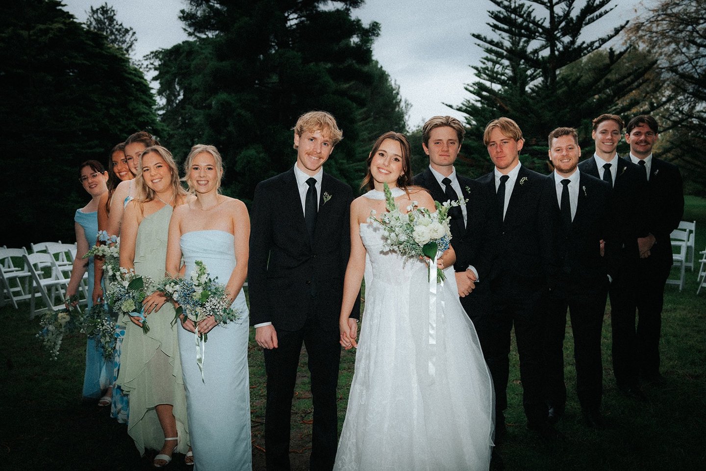 A bride and groom posing outdoors with their wedding party in light blue dresses and black suits.