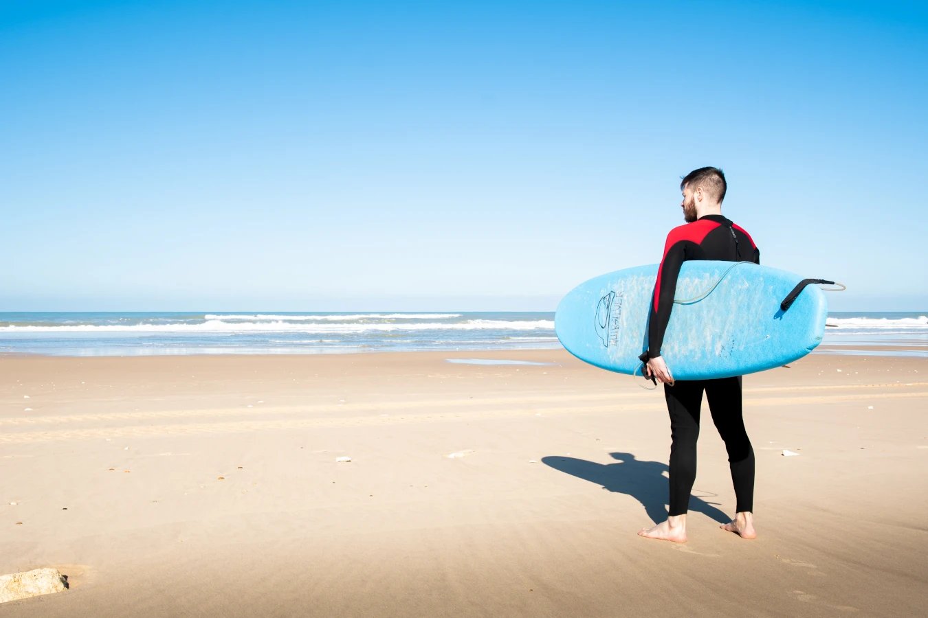 En homme en combinaison de surf, avec une planche bleu sur une plage