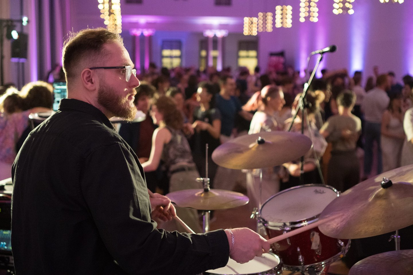 A bearded male drummer plays a live drum set at a crowded indoor party with purple lighting.