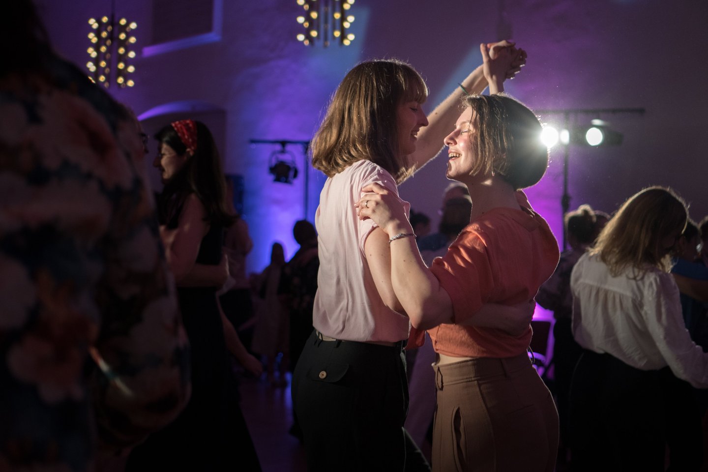 Two women smiling and dancing together at a lively party with purple event lighting.