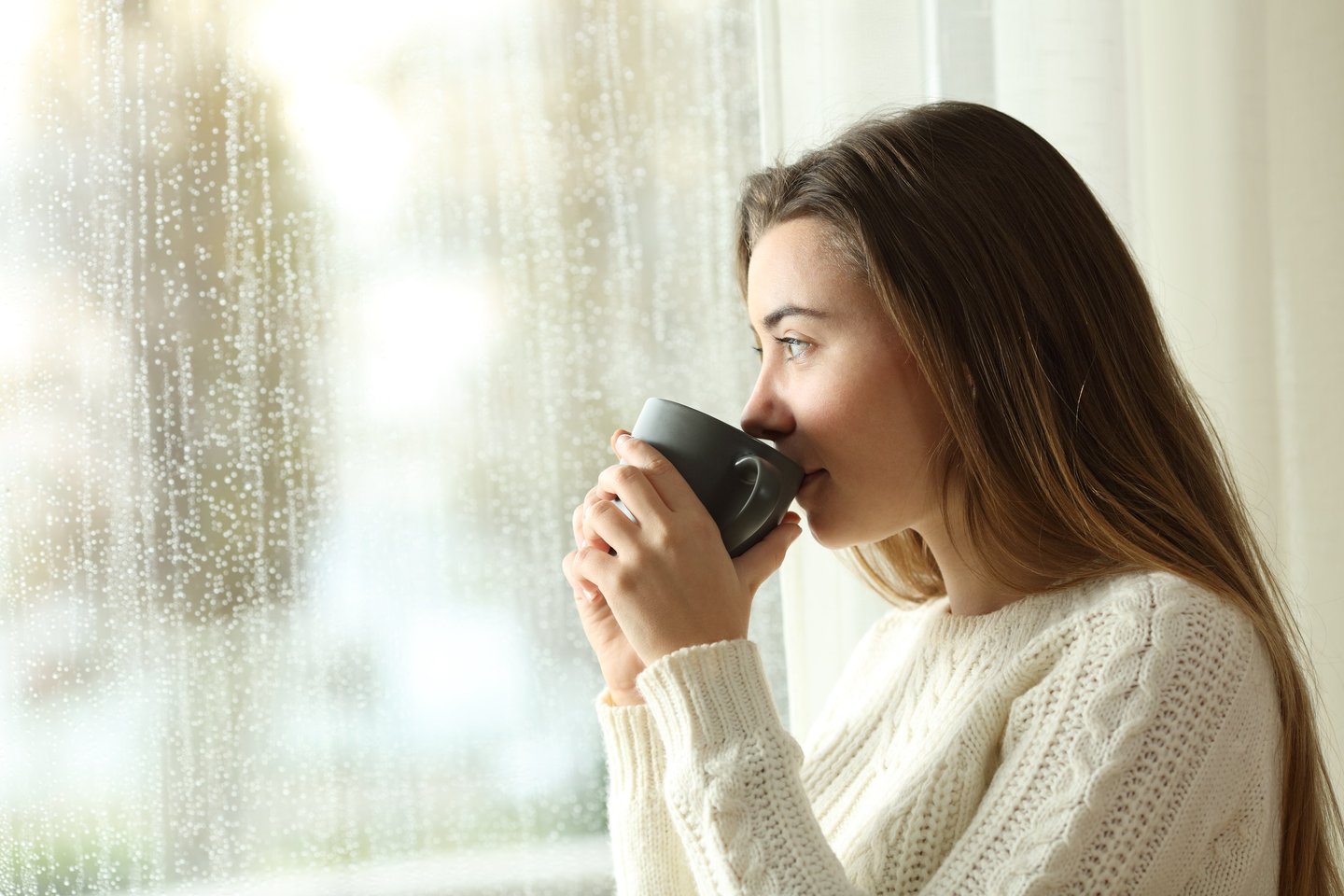 a woman drinking from a mug sitting by a window and its raining outisde