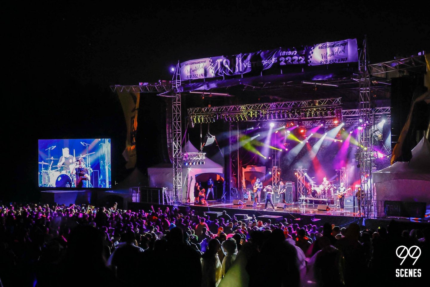 Rock band performing on an outdoor stage at night with colorful lights and a large crowd.