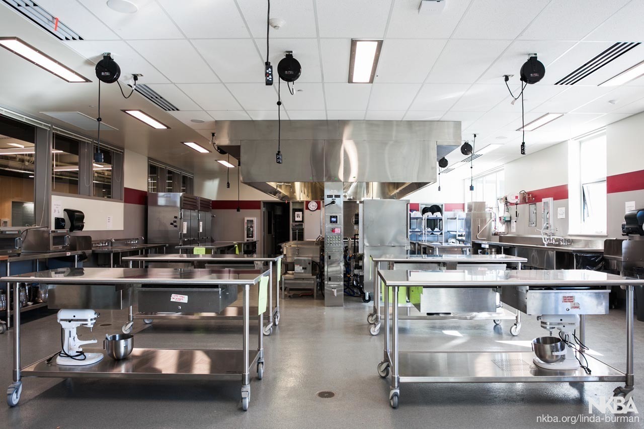 Wide shot of a professional kitchen with stainless steel workstations. Discover more about us.
