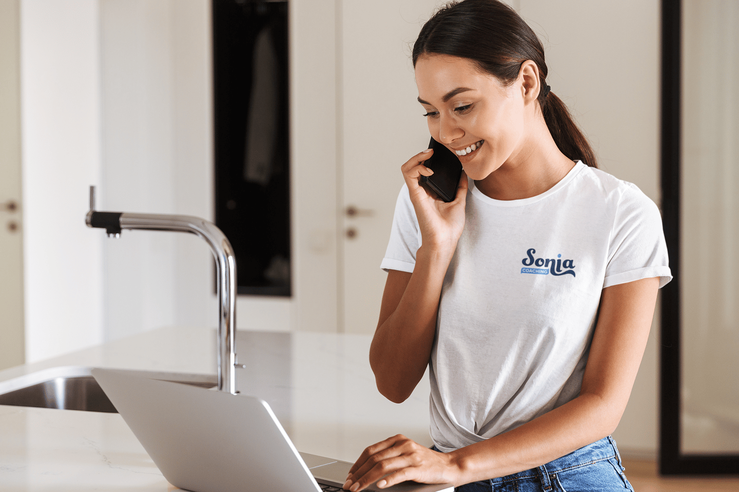 Femme souriante en t-shirt blanc avec un logo de coaching imprimé, parlant au téléphone.