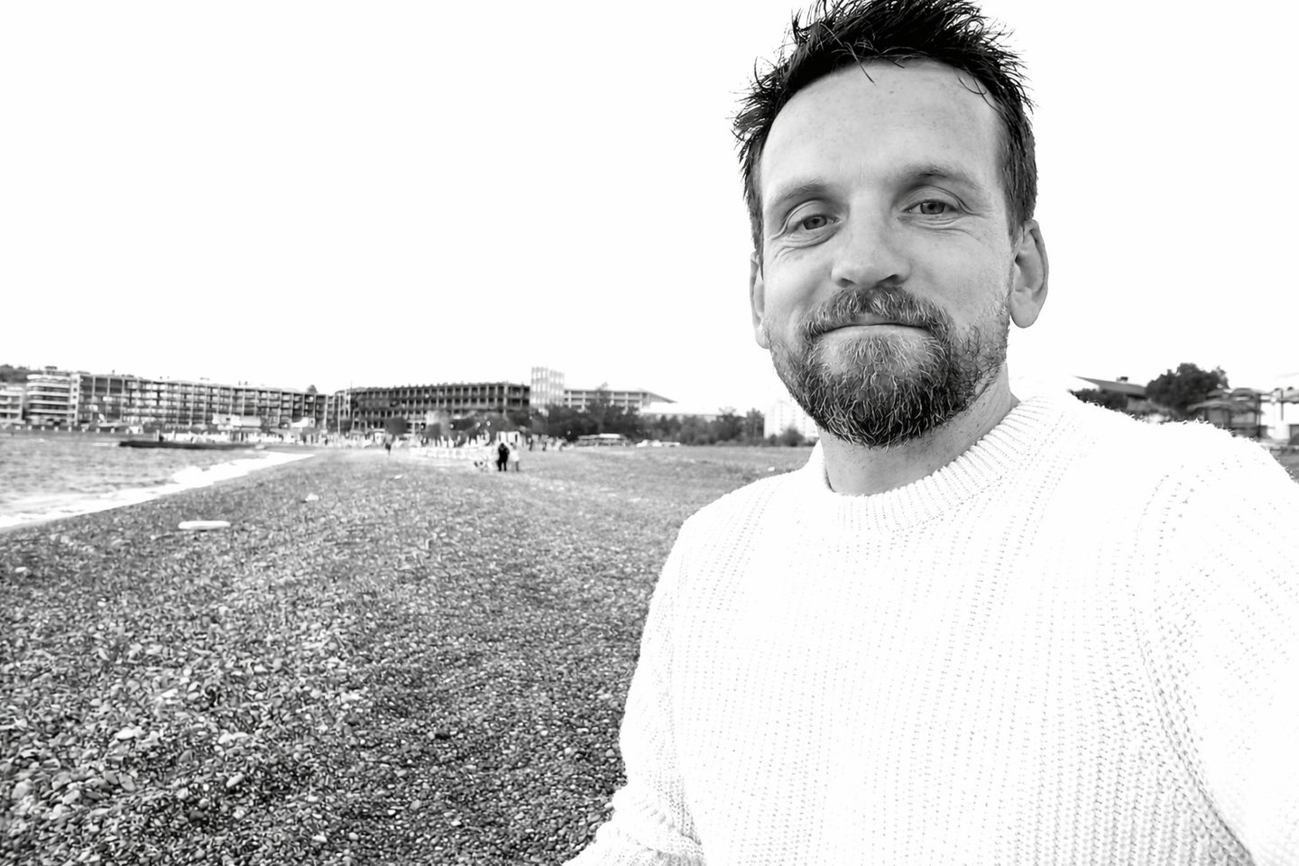A bearded man in a white sweater smiling on a rocky beach in a black and white portrait.