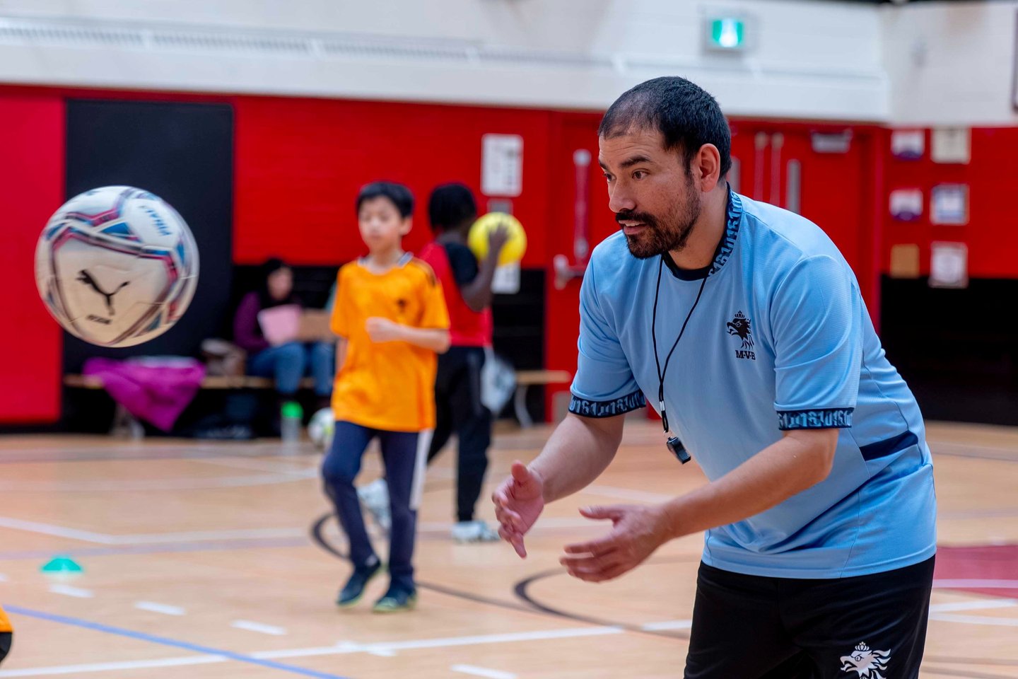 A youth soccer coach in a blue jersey training children during an indoor soccer sports class at MVB