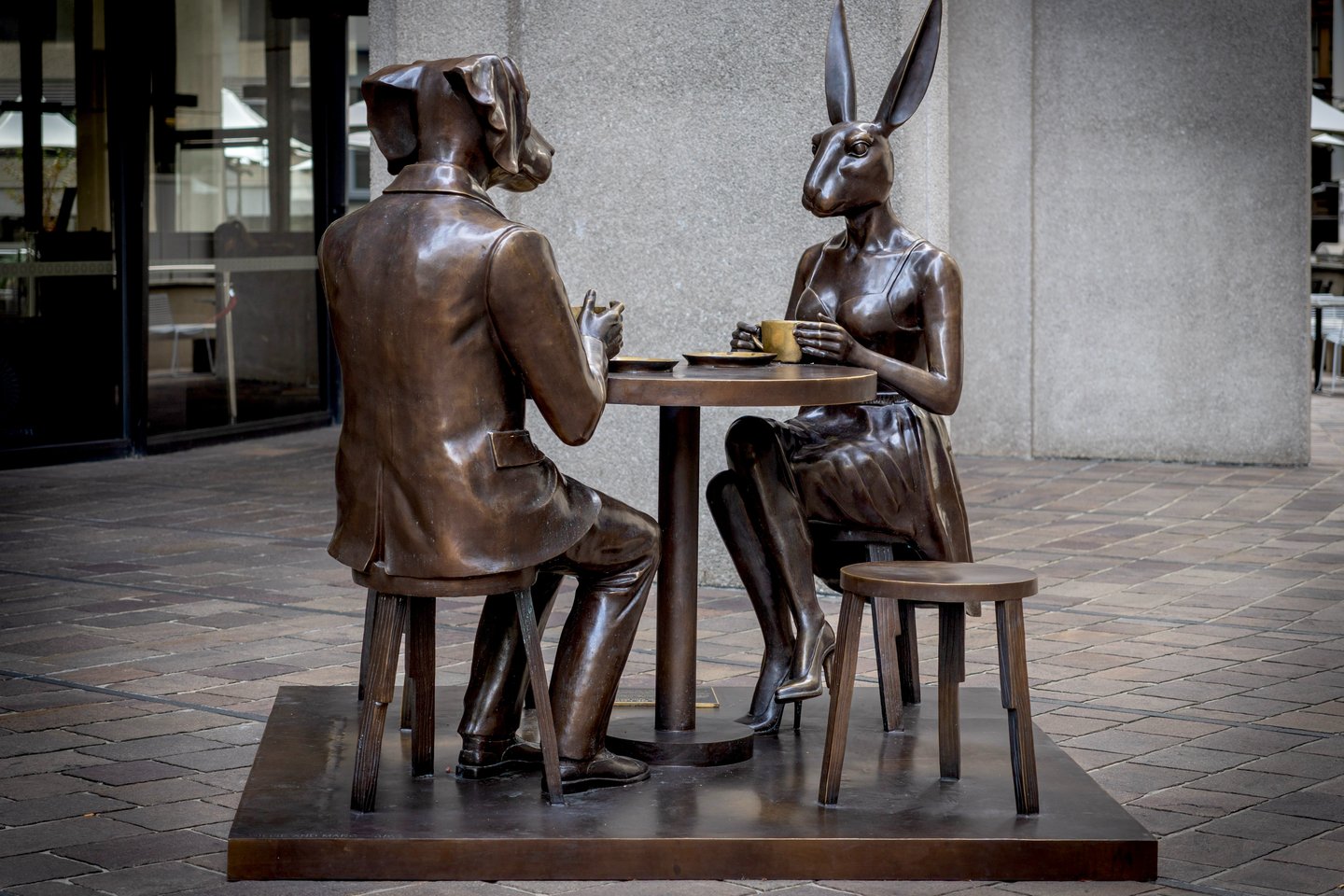 Bronze sculpture of a dog-headed man and rabbit-headed woman drinking coffee at a table.
