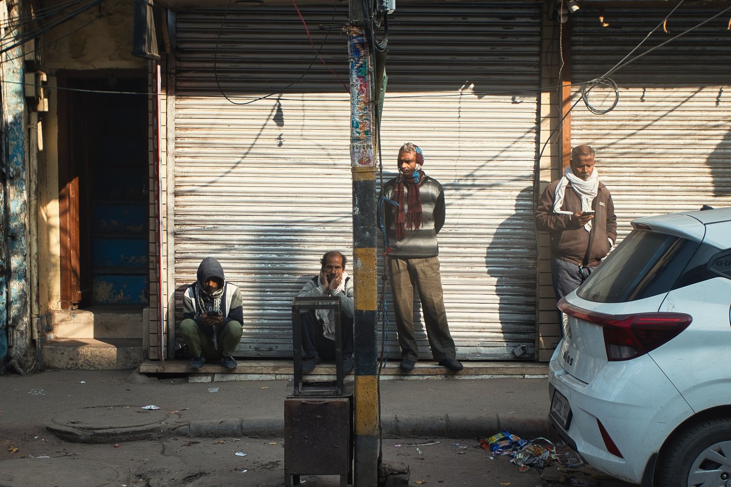 Men standing and sitting outside shuttered shops on a quiet Old Delhi street.