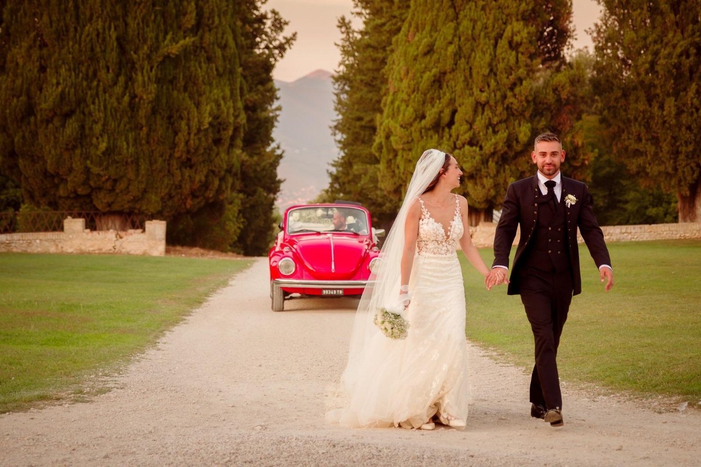 Sposi che camminano per mano lungo un viale alberato, sorridenti davanti alla loro auto d’epoca