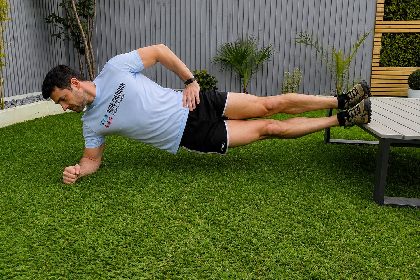 A client performing a side plank core exercise on green grass with feet elevated on a bench.