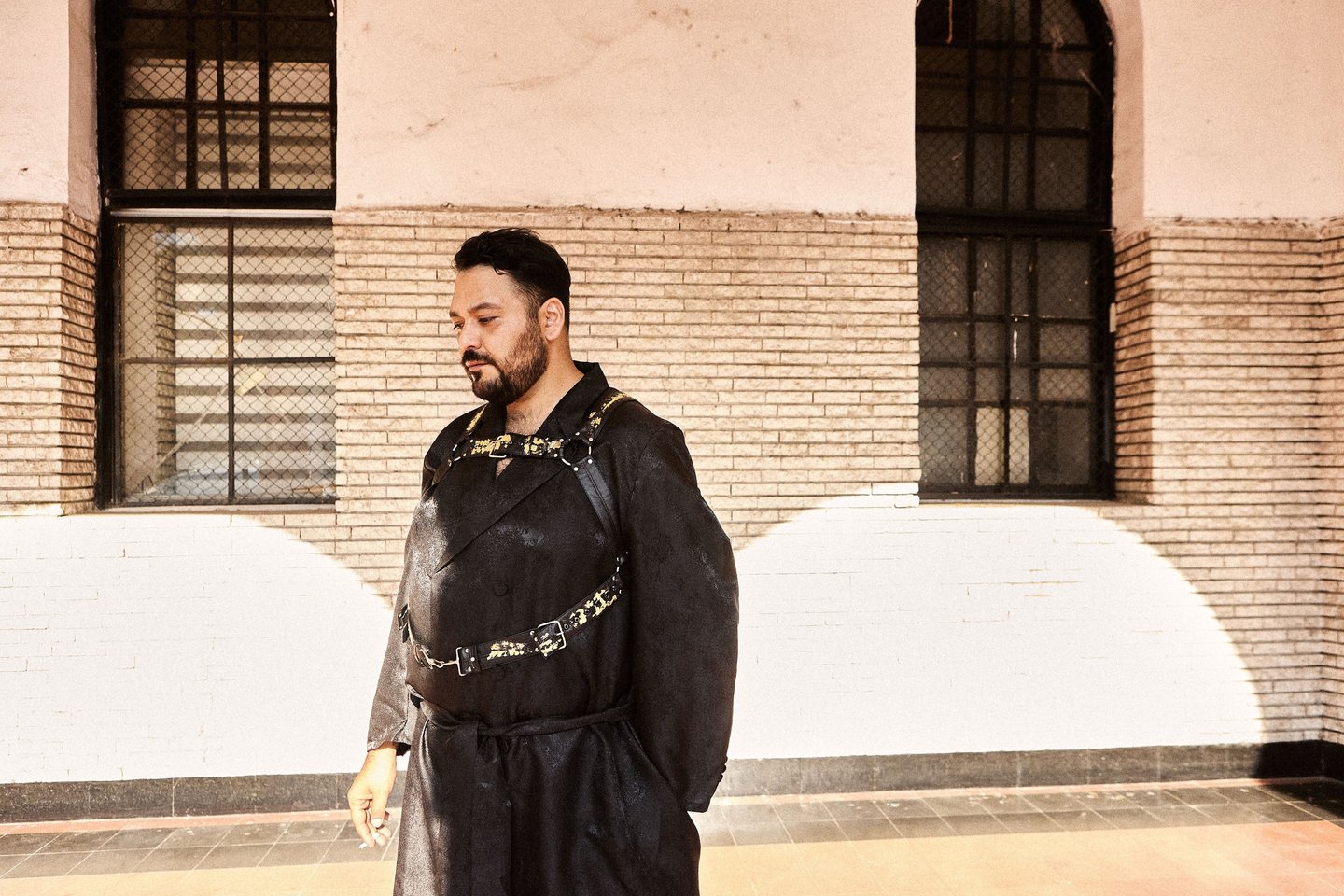 A man in a black avant-garde robe and leather body harness stands before a brick wall with windows.
