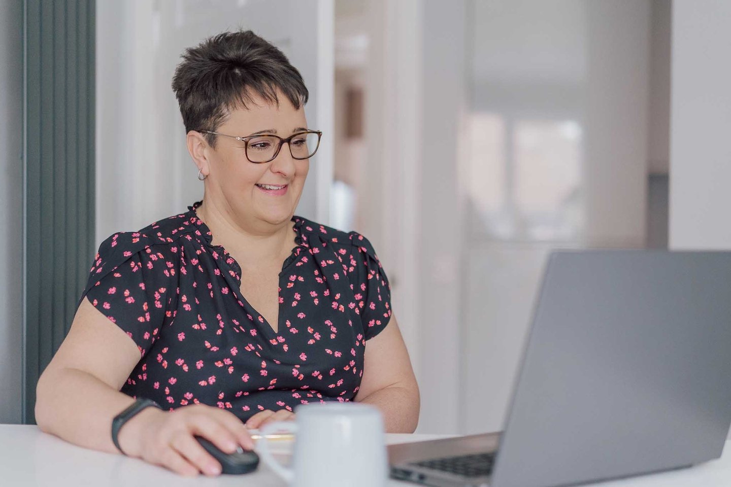 Smiling woman with short hair and glasses working on her laptop at a desk.