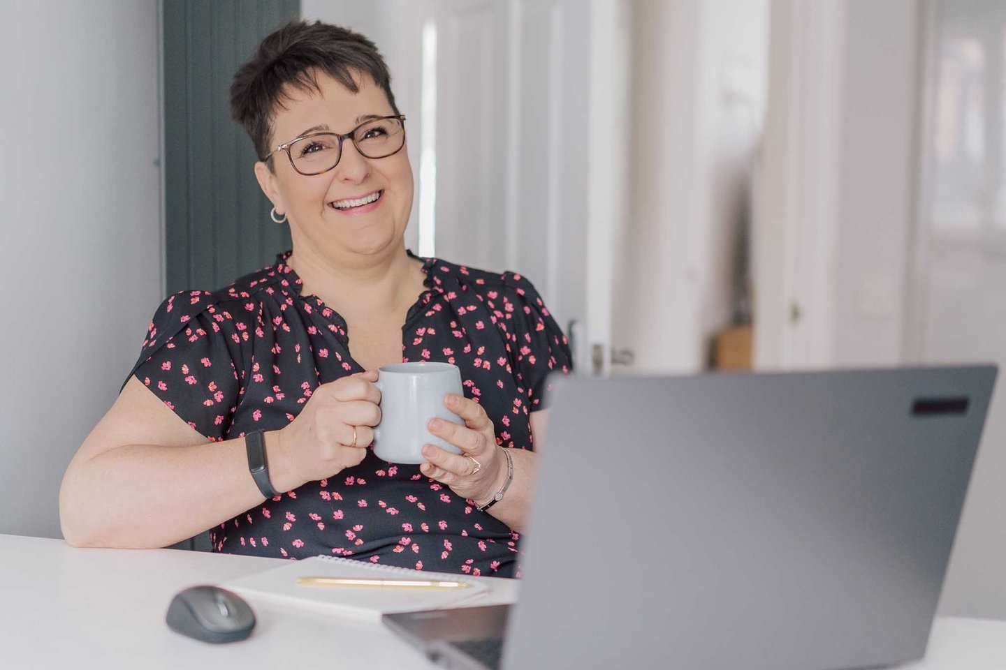 Smiling woman with short hair holding a coffee mug while working on her laptop at home.