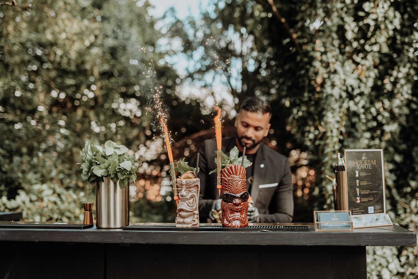 Professional bartender serving tropical tiki cocktails with sparkling garnishes at an outdoor garden bar.