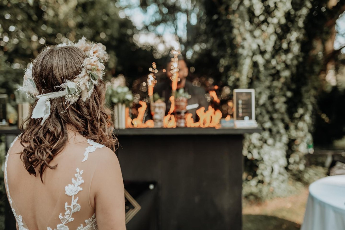 A bride with a floral crown watches a flair bartender perform a fire show at an outdoor wedding reception.