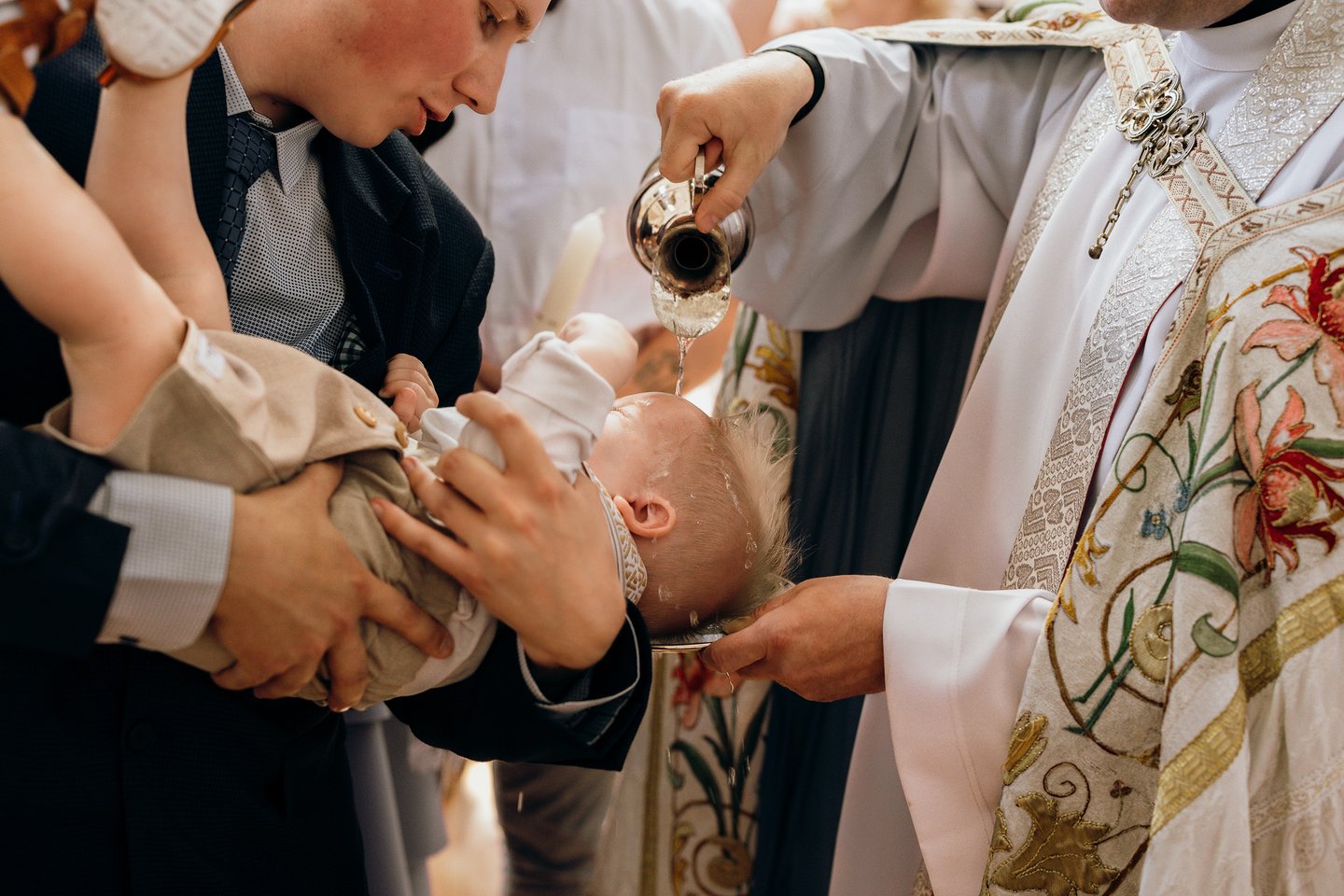 Krikštynų fotografija. Vyksta berniuko krikštas. Kunigas lieja vandenį ant vaiko kaktos.