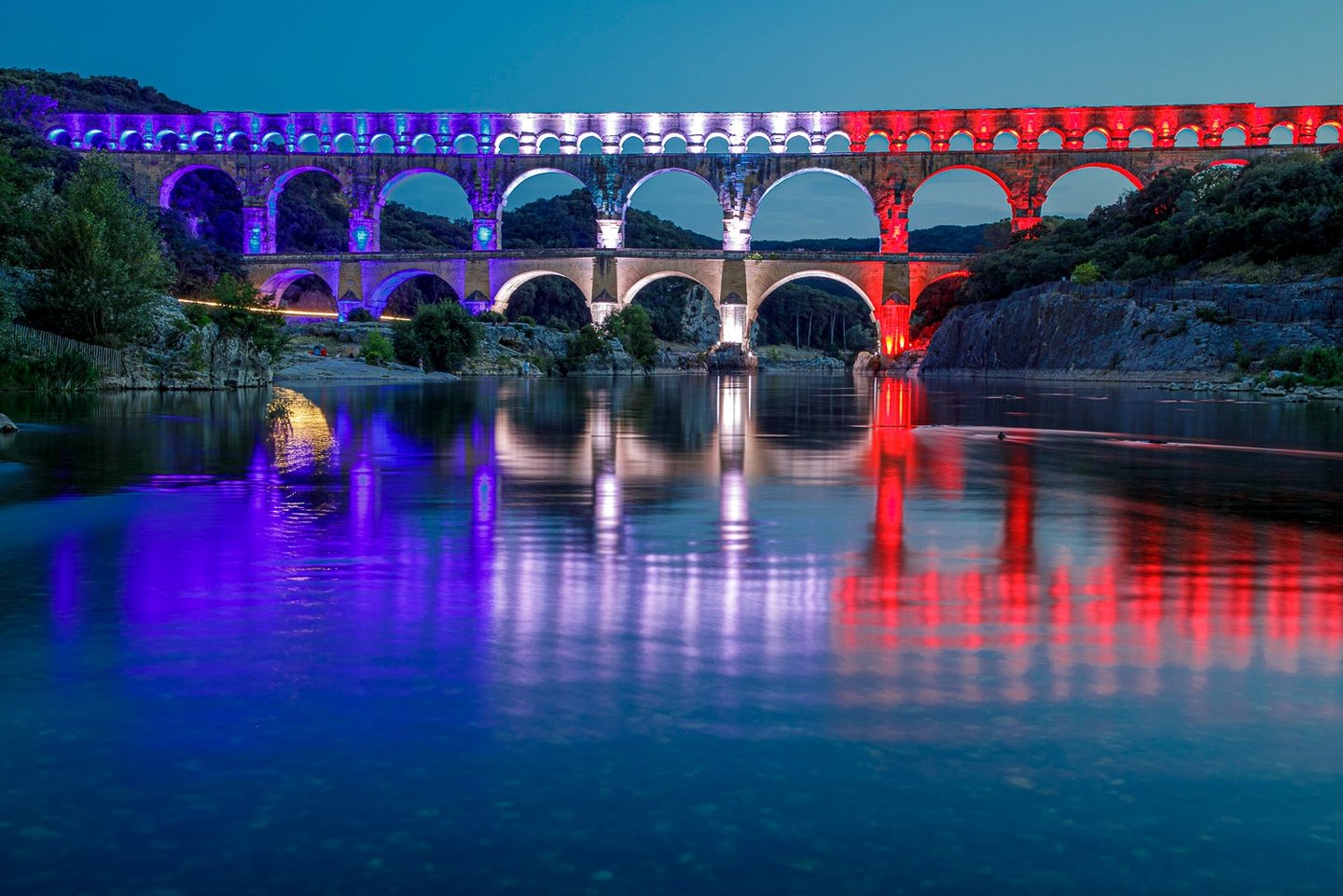 The Pont du Gard Roman aqueduct bridge illuminated in red, white, and blue lights reflecting on the Gardon River at night.