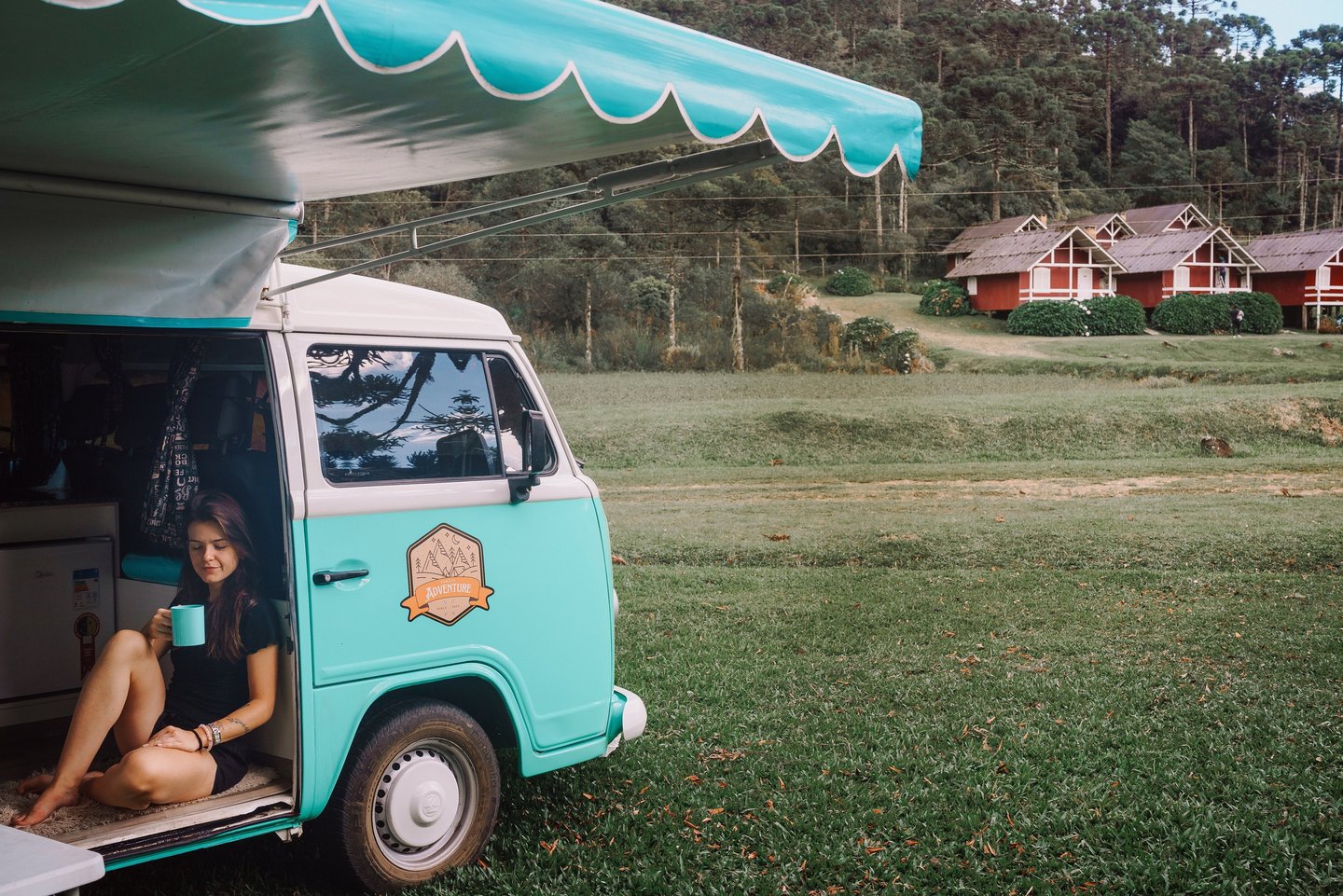 Uma pessoa sentada bebendo café dentro de uma kombi home, estacionada em um jardim.