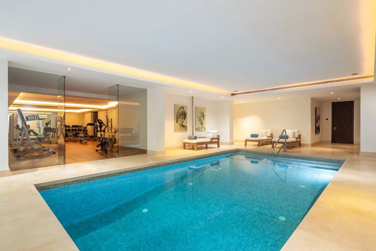 Luxury indoor swimming pool with lounge chairs and adjacent glass-walled fitness center.