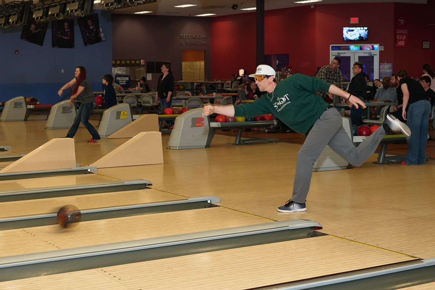 A male bowler in a green shirt releases a bowling ball down a wooden lane at a bowling alley.