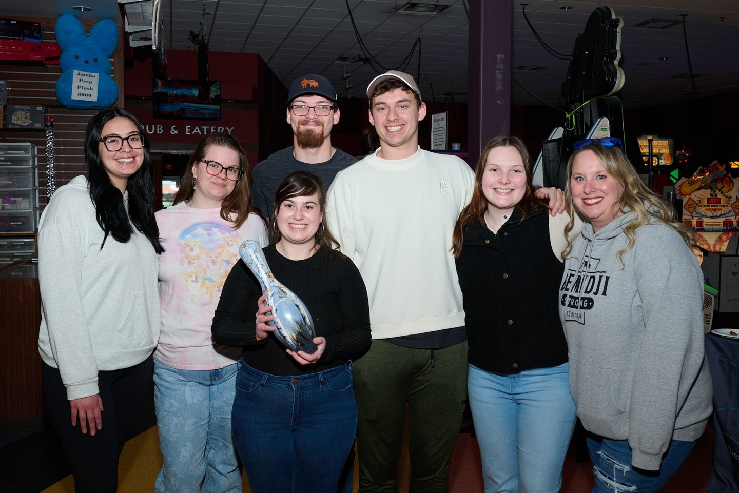 A group of smiling friends posing at a bowling alley while holding a decorated souvenir bowling pin.