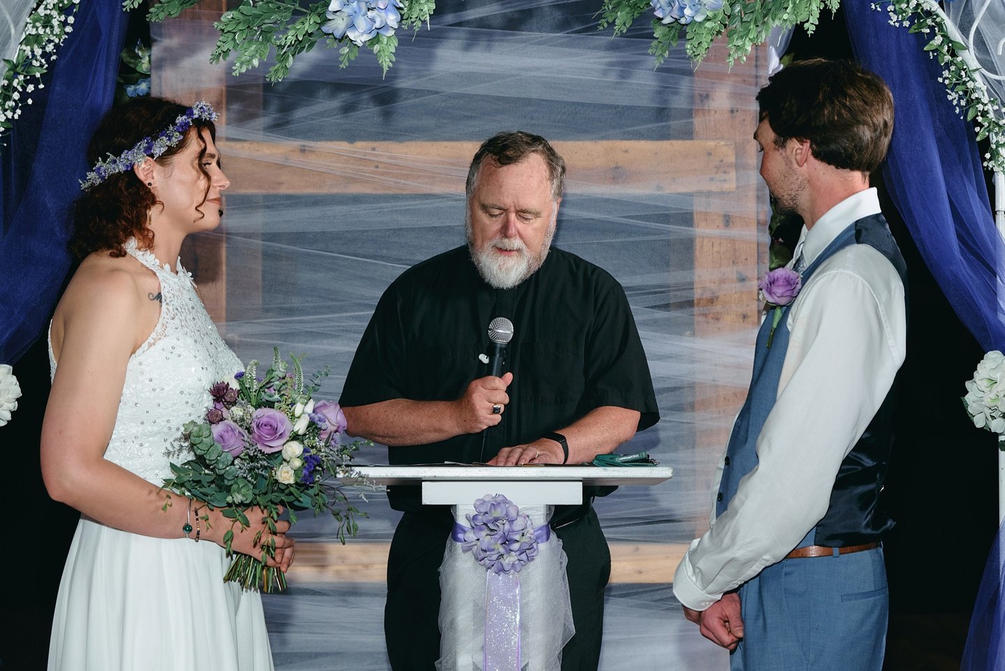a groom and a bride standing across each other and a priest standing in between them giving a speech