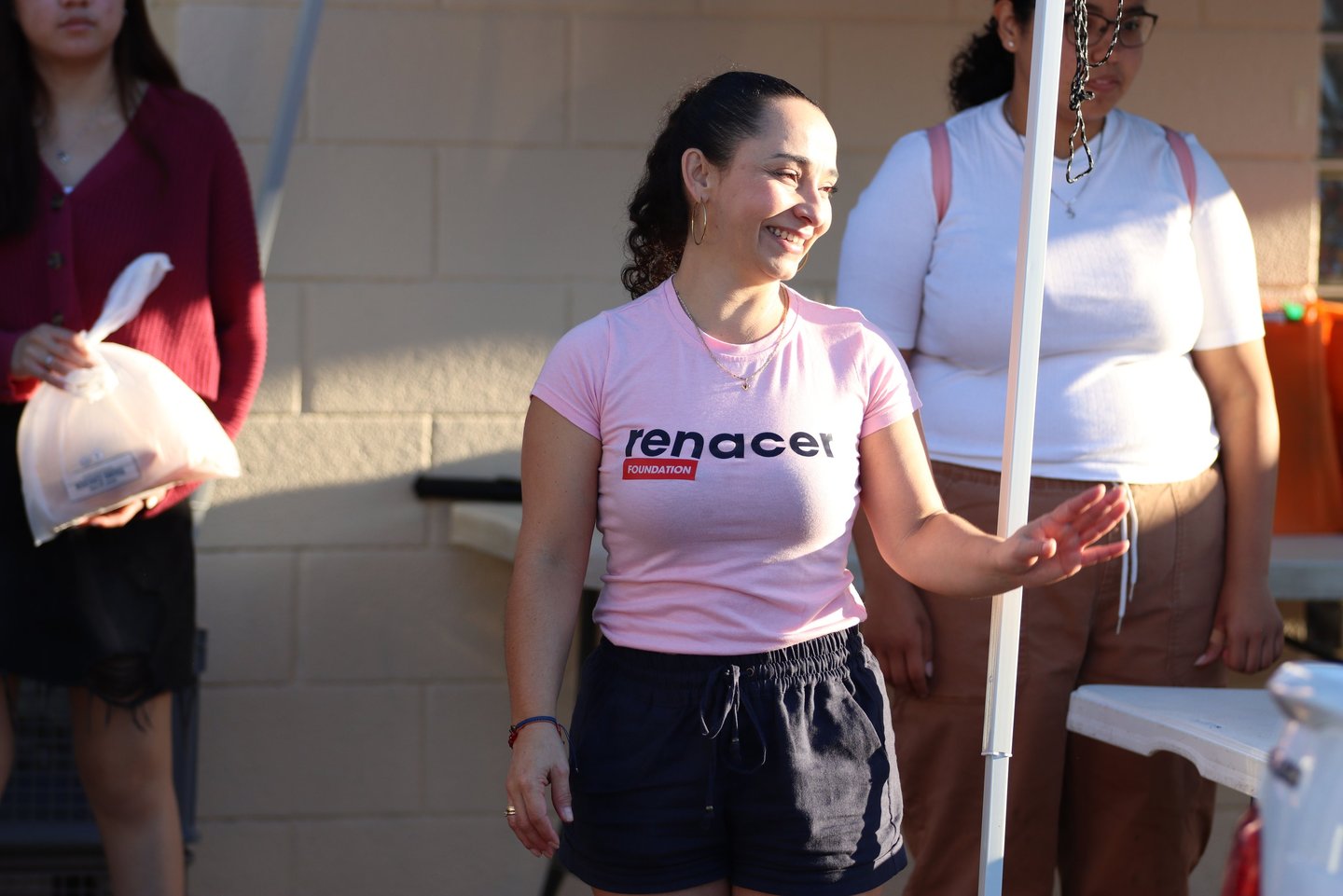 A smiling woman wearing a pink Renacer Foundation t-shirt volunteering at a community outreach event.