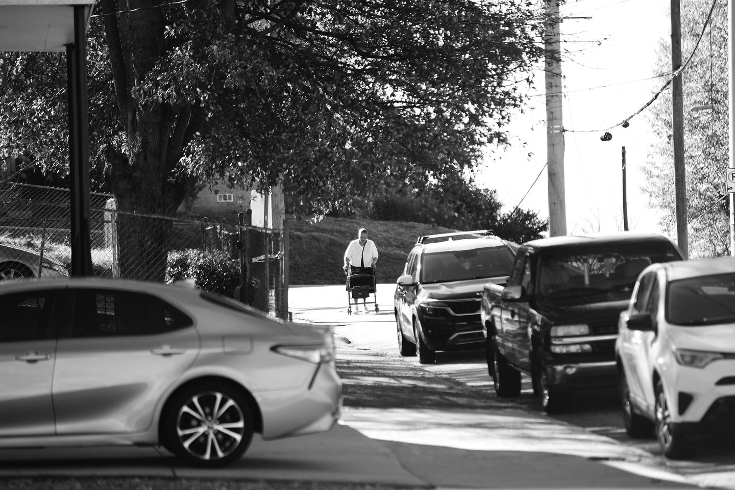 Black and white street photography of an elderly person using a mobility walker on a sidewalk lined with parked cars.