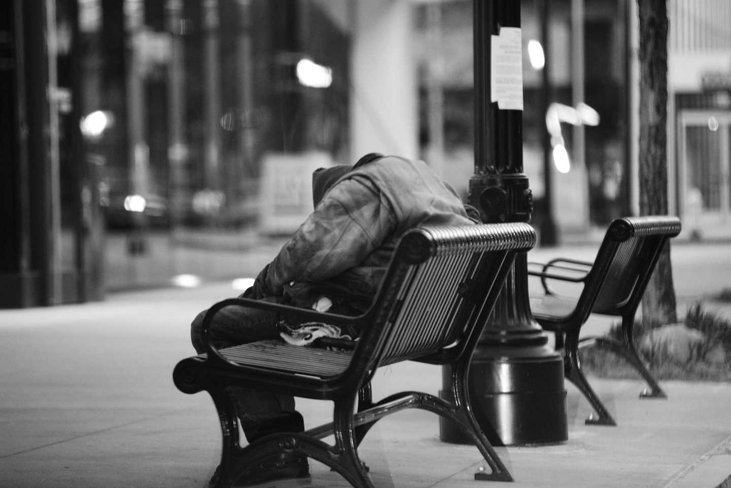 Black and white photo of a homeless person sleeping on a city park bench at night.