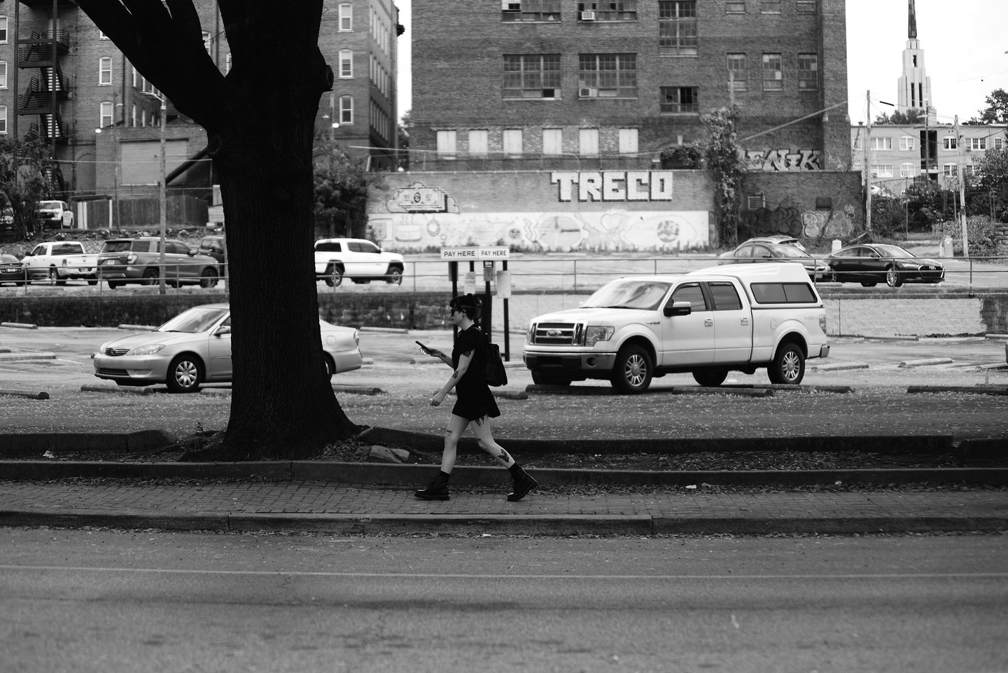 A person walks on a city sidewalk past parked cars, a large tree, and a building with "TRECO" painted on it, in black and