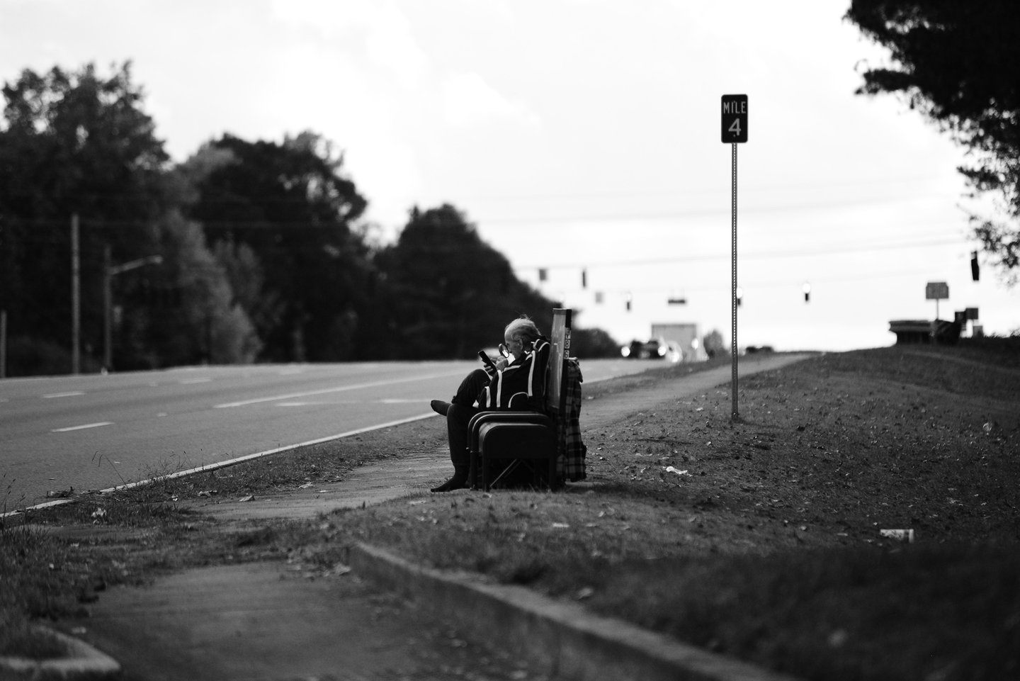 Black and white street photography of a person sitting on a bench by a highway using a smartphone.