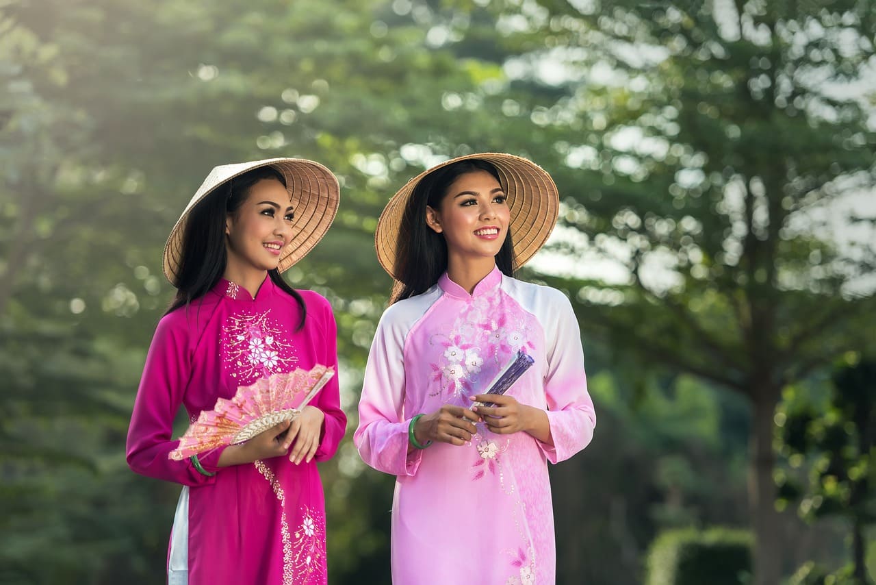 Zwei lächelnde Frauen in traditionellen vietnamesischen Ao Dai Kleidern mit kegelförmigen Strohhüten